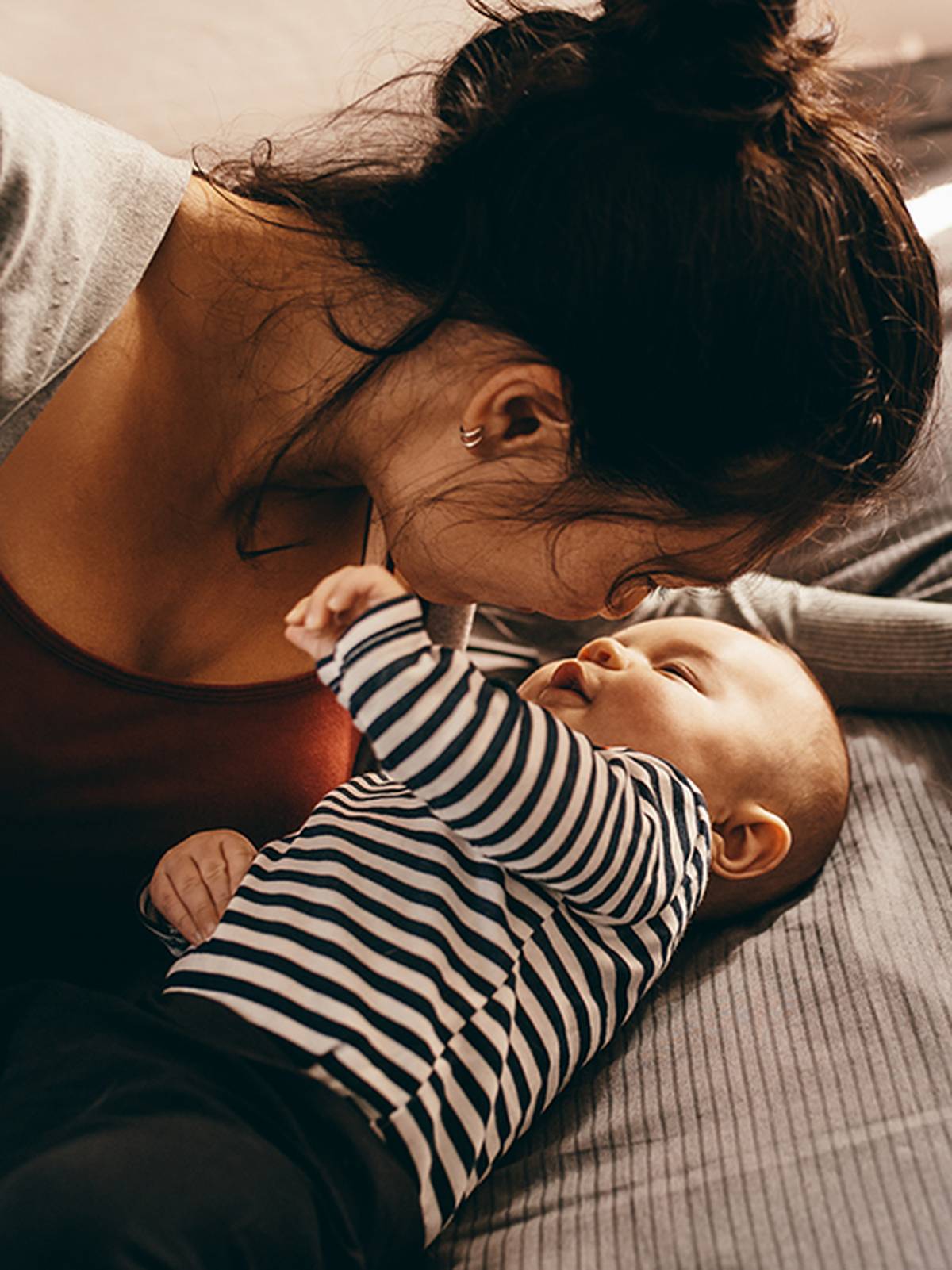 Eine Frau schmiegt liebevoll ihre Nase an ein Baby, das auf einem Bett liegt; beide wirken zufrieden und vertraut; das Baby trägt ein gestreiftes Hemd.