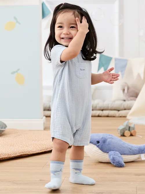 Ein lächelndes Kleinkind in einem hellblauen Strampler steht auf einem Holzboden, umgeben von Spielzeug und Dekoration, und streckt die Arme nach schwebenden Seifenblasen aus.