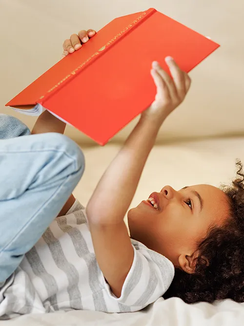 Ein Kind liegt vor hellem Hintergrund und liest vergnügt ein aufgeschlagenes rotes Buch. Es trägt ein gestreiftes Hemd und Jeans und hat einen fröhlichen Gesichtsausdruck.