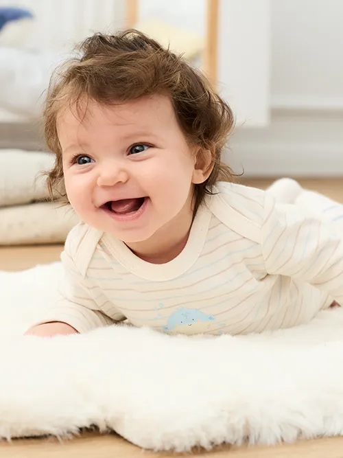 Ein lächelndes Baby mit lockigem Haar liegt in einem hellen Zimmer auf einem weichen weißen Teppich und trägt einen hell gestreiften Strampler.
