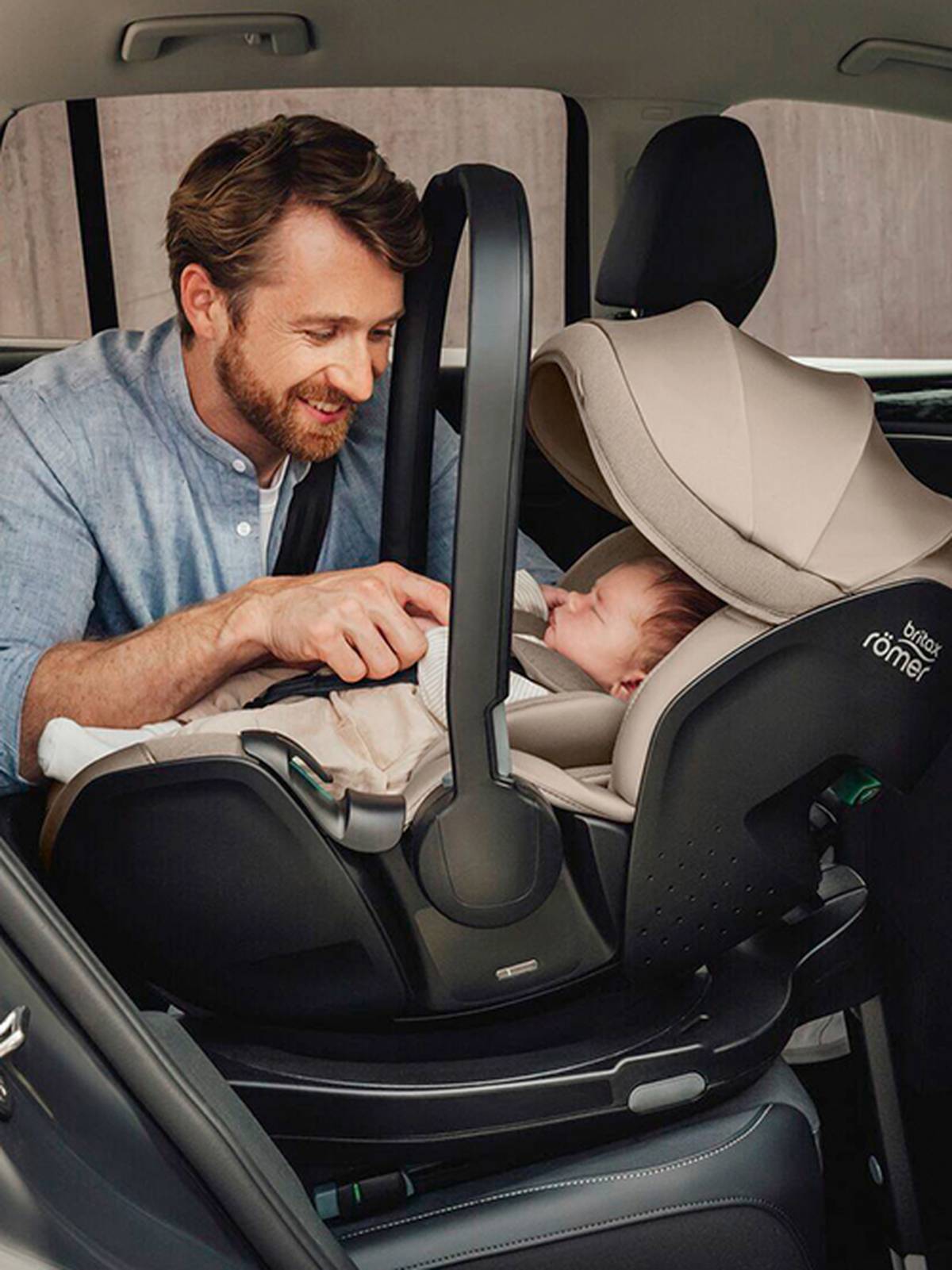 Ein Mann in einem blauen Hemd lächelt ein Baby in einem beigen Kindersitz in einem Auto an. Das Baby schläft friedlich.