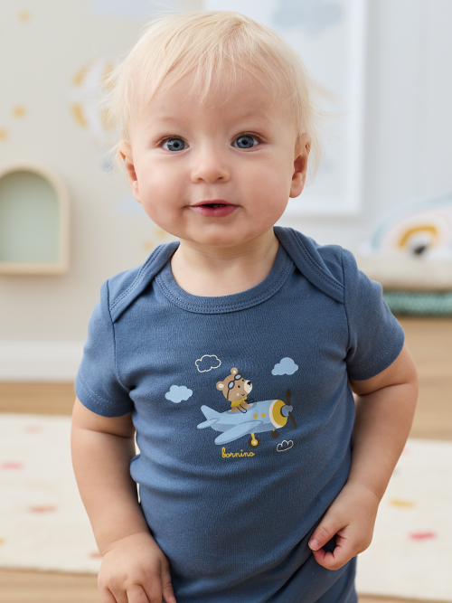 Kleinkind in einem blauen Strampler mit Bären- und Flugzeugmotiv, das drinnen steht und fröhlich dreinschaut.