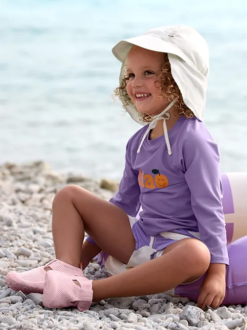 Kind in lila Kleidung und Sonnenhut sitzt lächelnd an einem Kieselstrand; im Hintergrund ist eine aufblasbare Luftmatratze zu sehen.