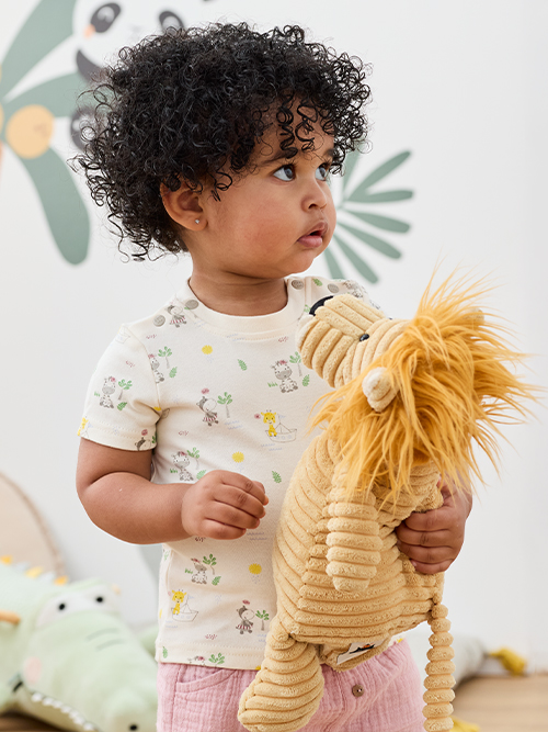 Ein Kleinkind mit lockigem Haar, das ein gemustertes Hemd trägt, hält ein Plüschtier in Form eines Löwen in der Hand. Im Hintergrund ist eine Wandtapete mit Blattmuster zu sehen.