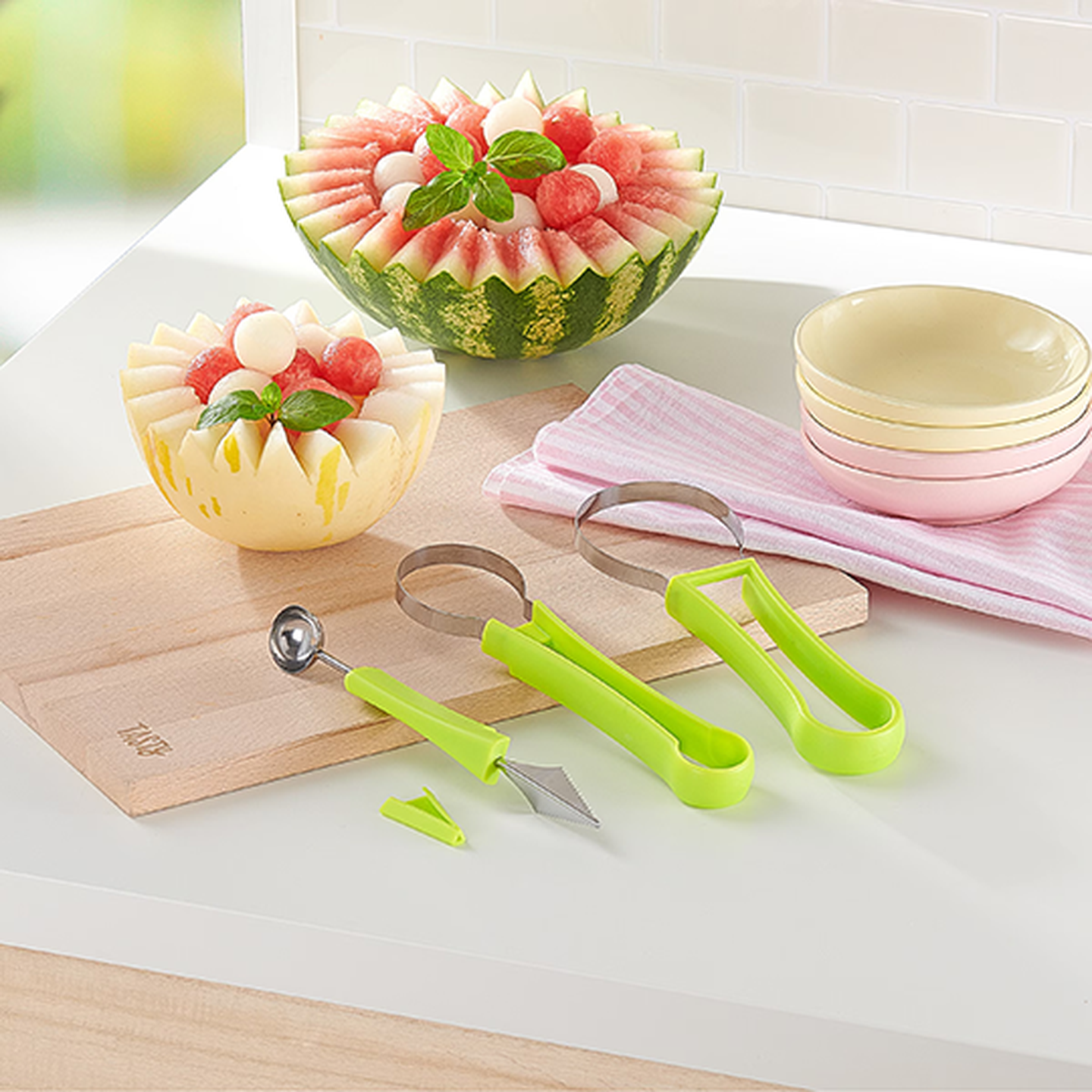Zwei Melonenschalen, gefüllt mit Wassermelonen- und Melonenbällchen, umgeben von grünen Küchenutensilien auf einem Holzbrett, mit gestapelten Schalen und einem Tuch.