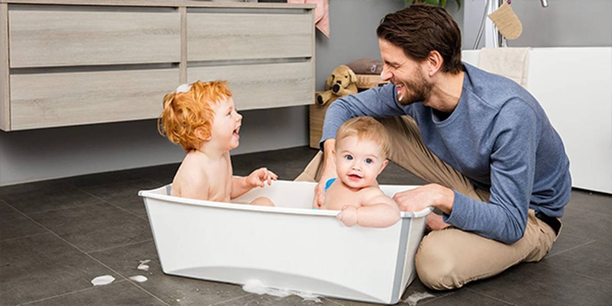 Ein Vater beobachtet freudig zwei Kinder, die in einer weißen Badewanne auf dem gefliesten Badezimmerboden spielen, umgeben von Badespielzeug und Seifenblasen.