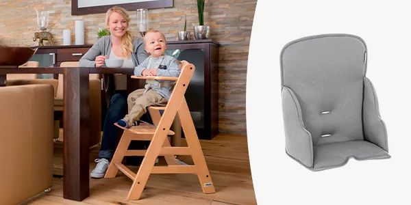 Ein Baby sitzt in einem hölzernen Hochstuhl an einem Esstisch neben einer lächelnden Frau. Daneben liegt ein separates, gepolstertes graues Kissen.