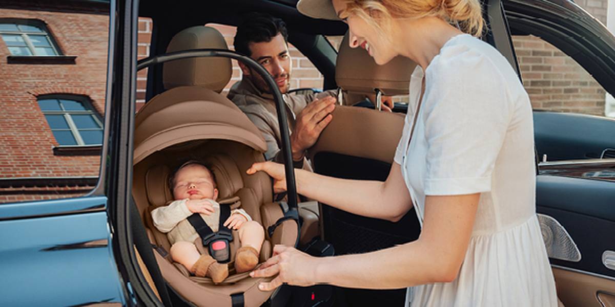 Eine Frau setzt ein Baby in einen Autositz, während ein Mann zusieht. Das Baby schläft friedlich. Die Szene spielt neben einem geparkten Auto in der Nähe eines Backsteingebäudes.