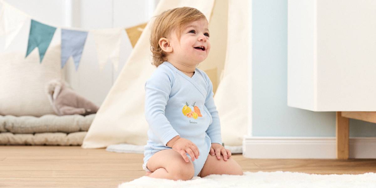 Ein fröhliches Kleinkind in einem hellblauen Strampler kniet auf einem weichen weißen Teppich, umgeben von bunten Wimpelketten und Kissen in einem gemütlichen, lichtdurchfluteten Zimmer.