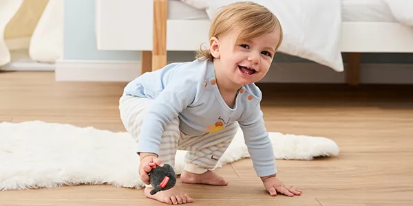 Ein lächelndes Kleinkind in gestreiftem Schlafanzug hockt auf einem Holzboden und hält eine kleine Spielzeugmaus in der Hand; im Hintergrund ist ein gemütliches Schlafzimmer zu sehen.