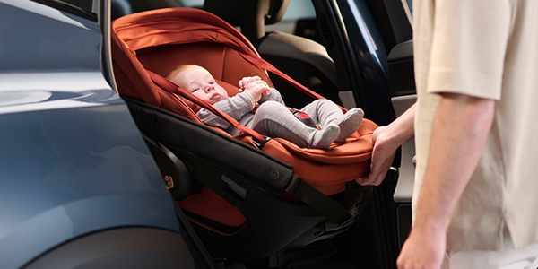 Ein Baby in einem orangefarbenen Autositz wird von einem Erwachsenen behutsam aus einem Auto gehoben.