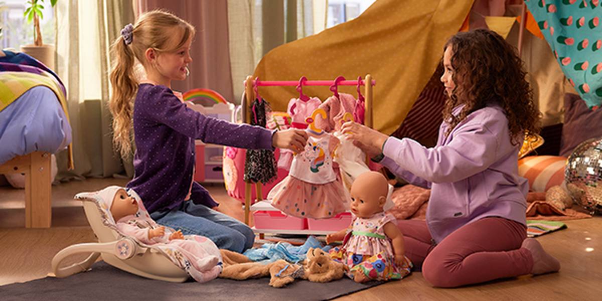 Zwei Mädchen spielen in einem gemütlichen Zimmer mit Puppen und Kleidern. Eine Puppe sitzt in einer Babytrage, und in der Nähe steht ein Ständer mit bunten Outfits.