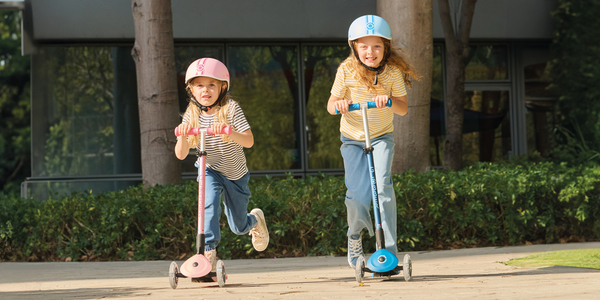Zwei Kinder mit Helmen fahren auf einem Weg Roller, umgeben von Grün und Bäumen, mit einem Gebäude im Hintergrund.