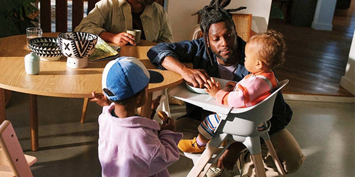 Ein Mann füttert ein Baby in einem Hochstuhl, während ein anderes Kind daneben steht; im Hintergrund sitzt eine Person an einem Holztisch mit gemusterten Schüsseln.