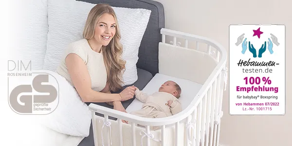 Eine Frau lächelt ein Baby an, das in einem Gitterbett neben einem Bett liegt; auf dem Bild ist ein Qualitätssiegel zu sehen.
