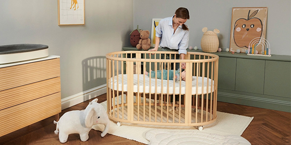 Eine Frau kümmert sich um ein Baby in einem hölzernen Kinderbett in einem gemütlichen Kinderzimmer mit Spielzeug und Dekoration, darunter ein Plüschelefant und Kunstwerke an den Wänden.