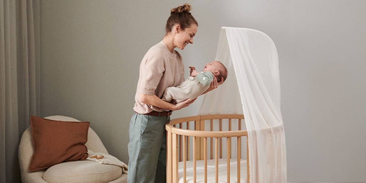 Eine Frau lächelt ein in eine Decke gehülltes Baby an; sie steht neben einer hölzernen Wiege mit einem durchscheinenden Betthimmel in einem sanft beleuchteten Kinderzimmer.