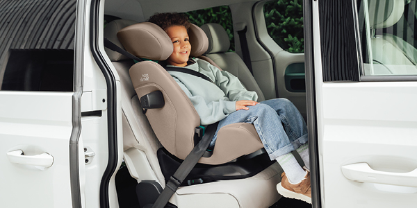 Ein lächelndes Kind sitzt sicher in einem Kindersitz in einem weißen Auto – dessen Tür geöffnet ist –, angeschnallt und in legerer Kleidung sowie Turnschuhen.