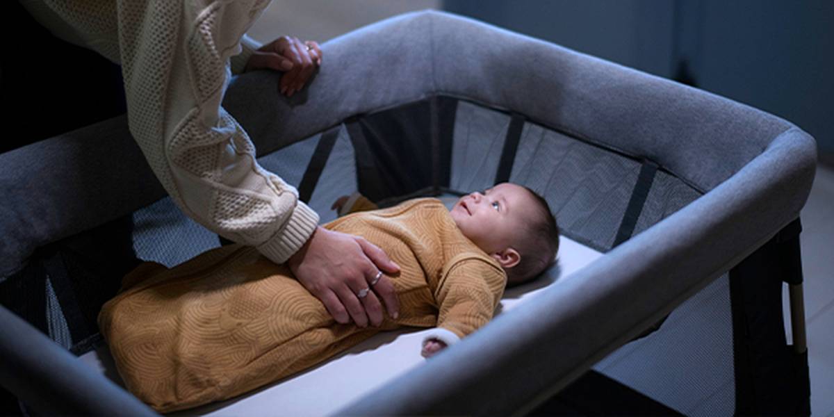 Ein Erwachsener bettet ein Kleinkind in ein graues Gitterbett. Das Baby trägt einen braunen Schlafsack und blickt nach oben.