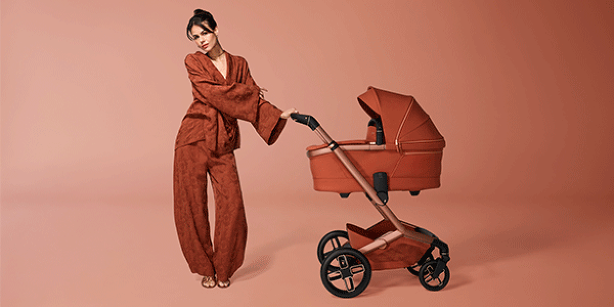 Person in rostfarbener Kleidung steht neben einem passenden Kinderwagen mit Babywanne vor einem einfarbig roséfarbenen Hintergrund. Kinderwagen und Outfit sind farblich aufeinander abgestimmt.