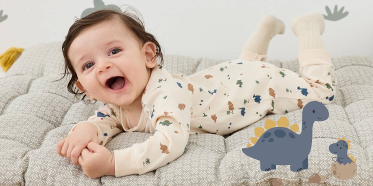 Baby liegt auf einer gepolsterten Spielmatte und trägt einen hellen Langarm-Overall mit Dinosauriermuster. Rechts neben dem Baby sind illustrierte Dinosaurier und ein schlüpfendes Dino-Baby dargestellt.