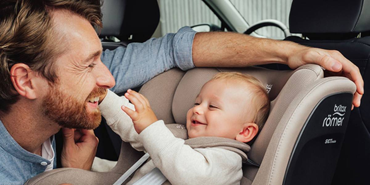 Ein Mann lächelt ein Baby an, das in einem Autositz im Wagen sitzt. Das Baby streckt die Hände nach dem Gesicht des Mannes aus und lächelt fröhlich.