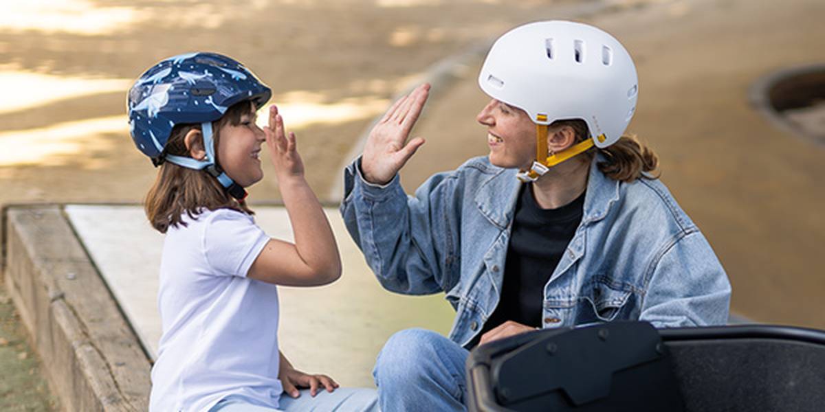 Ein Erwachsener und ein Kind, die beide Helme tragen, geben sich ein High-Five und lächeln sich an, während sie an einem sonnigen Tag draußen sitzen.