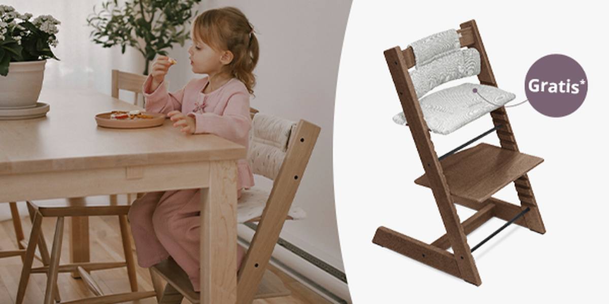 Ein junges Mädchen sitzt an einem Esstisch und isst. Neben ihm steht ein leerer Hochstuhl aus Holz mit Kissen. Auf dem Etikett ist ein „Gratis“-Schild zu sehen.