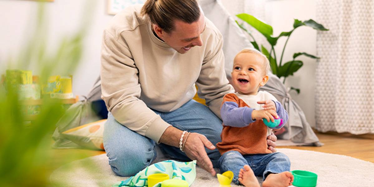 Ein lächelnder Mann und ein Baby sitzen auf einem Teppich und spielen mit Spielzeug in einem hellen Raum. Im Hintergrund sorgen grüne Pflanzen für eine freundliche und gemütliche Atmosphäre.