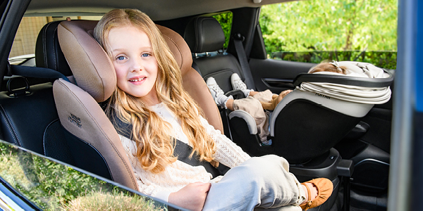 Ein junges Mädchen sitzt lächelnd in einem Autositz, während neben ihm in einem Fahrzeug ein Baby in einer Babyschale schläft.