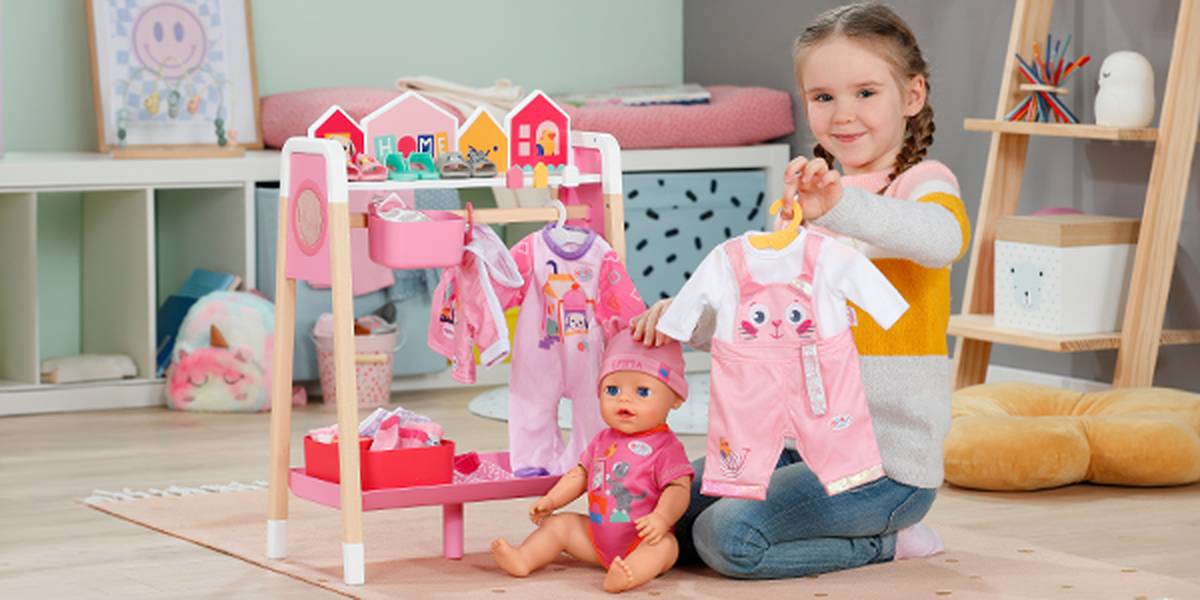 A young girl with braided hair plays in a room, sitting on the floor with a doll and a play clothes rack with colorful outfits.