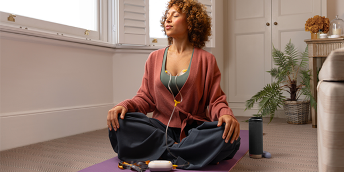 A woman sits cross-legged on a yoga mat, eyes closed in meditation. She's wearing earphones and a cozy cardigan, with a water bottle beside her.