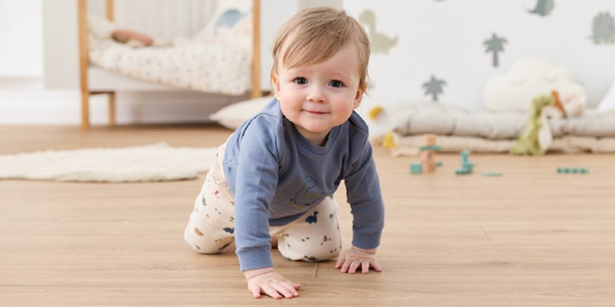 Ein Baby in einem blauen Hemd und einer gemusterten Hose krabbelt auf einem Holzboden in einem Kinderzimmer; im Hintergrund sind Spielzeug und ein kleines Bett zu sehen.