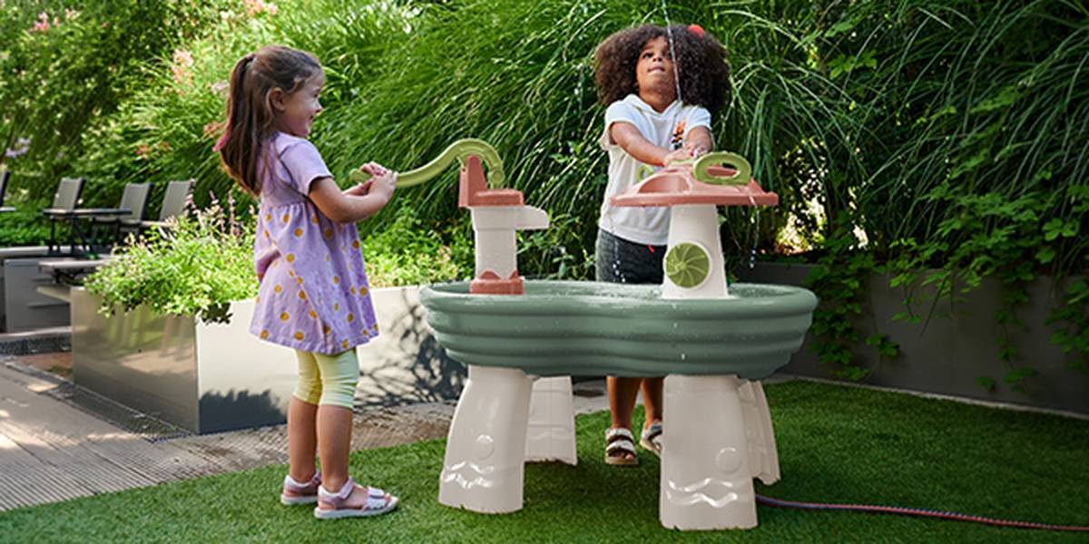 Zwei Kinder spielen im Freien an einem Wassertisch, umgeben von Grün. Der Junge und das Mädchen wirken vertieft in ihr Spiel und glücklich in der Gartenumgebung.