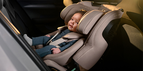 Ein Baby in blauer Kleidung sitzt sicher in einem beigen Kindersitz, der rückwärtsgerichtet auf der Rückbank eines Autos angebracht ist.