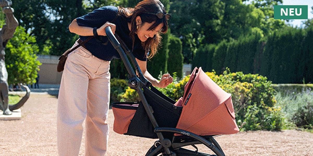 A woman leans over a stroller with a pink canopy, smiling, surrounded by greenery in a park setting.