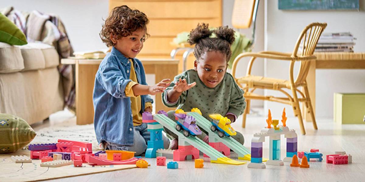 Zwei kleine Kinder spielen in einem hellen Wohnzimmer mit bunten Bauklötzen und Spielzeugautos; im Hintergrund sind ein Sofa und ein Sessel zu sehen.