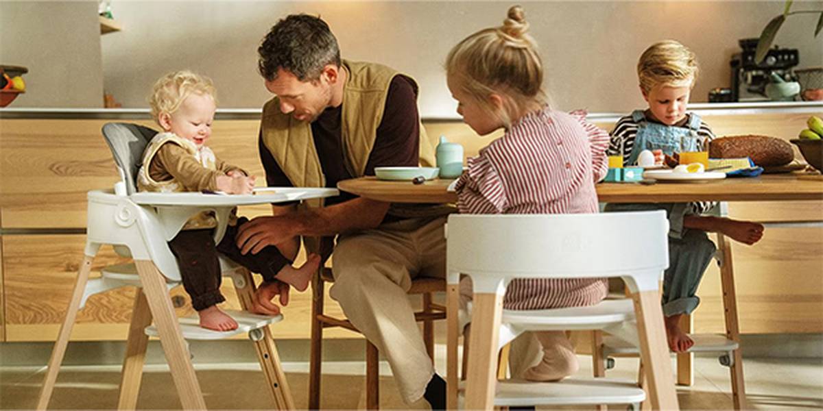Eine vierköpfige Familie genießt eine Mahlzeit in einer modernen Küche. Der Vater unterhält sich mit einem Kleinkind im Hochstuhl, während zwei Kinder am Tisch sitzen und essen.