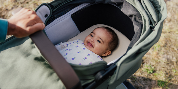 Baby liegt in der grünen Babywanne eines Kinderwagens. Die Babywanne wird von einer Hand am Tragebügel gehalten.