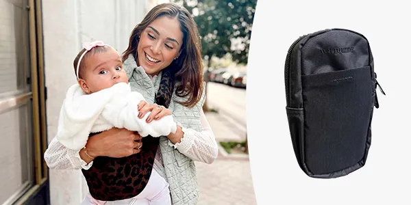 Frau trägt ein Baby in einem Tuch mit Leopardenmuster und lächelt im Freien; daneben liegt eine schwarze Tasche mit Reißverschluss.