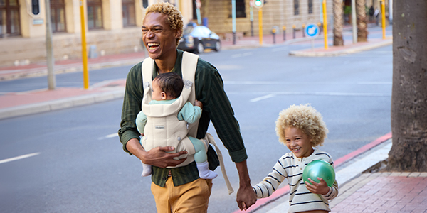 A man smiling while walking down a city street, carrying a baby in a front carrier and holding hands with a young child holding a green ball.