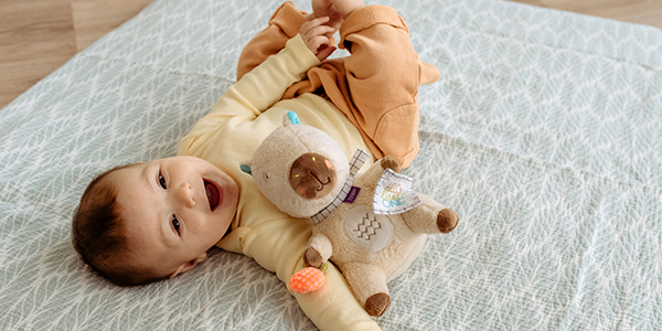 Ein Baby liegt auf einer gemusterten Decke, trägt ein gelb-orangefarbenes Outfit, hält ein Plüschtier und lächelt.