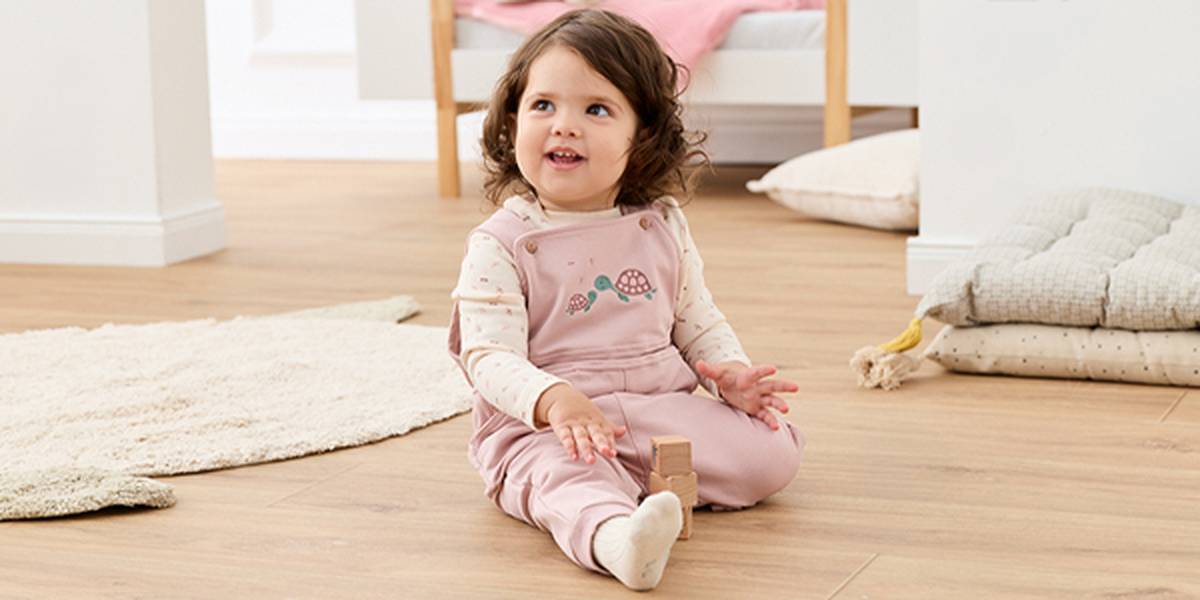 Ein Kleinkind in einem rosa Overall sitzt mit einem Holzklotz auf dem Boden, blickt nach oben und lächelt in einem gemütlichen Zimmer mit weichen Möbeln.