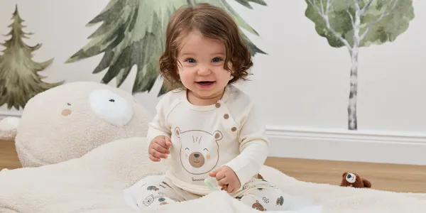 Ein lächelndes Baby mit lockigem Haar, bekleidet mit einem cremefarbenen Schlafanzug im Bären-Design, sitzt auf einer weichen, beigen Decke; im Hintergrund sind Plüschtiere und ein Baummotiv zu sehen.