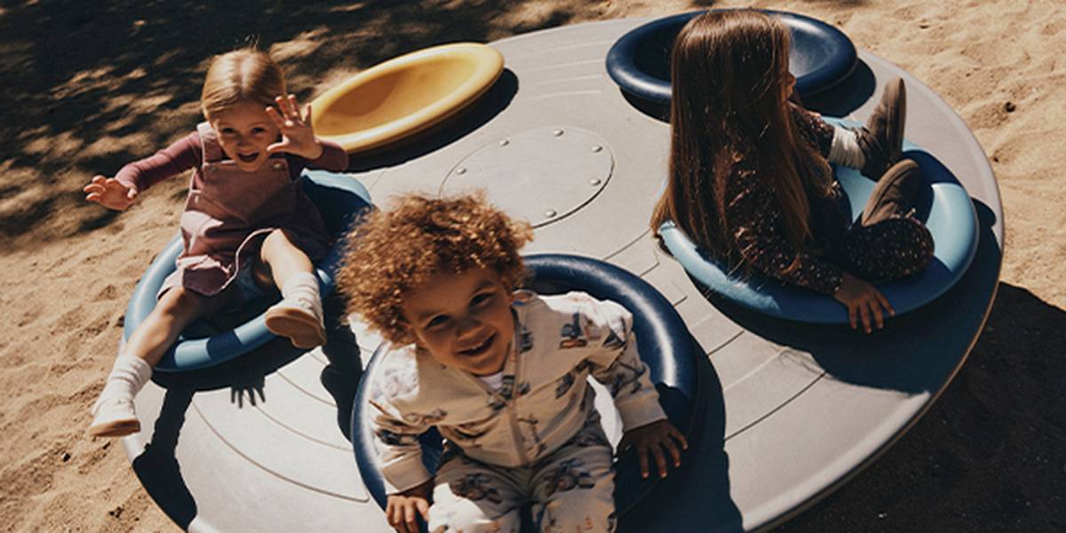 Drei Kinder sitzen auf einem großen Spielplatz-Karussell mit runden Sitzelementen in Blau und Gelb. Das Karussell steht auf einer Sandfläche im Freien und lädt zum gemeinsamen Spielen, Drehen und Entdecken ein.