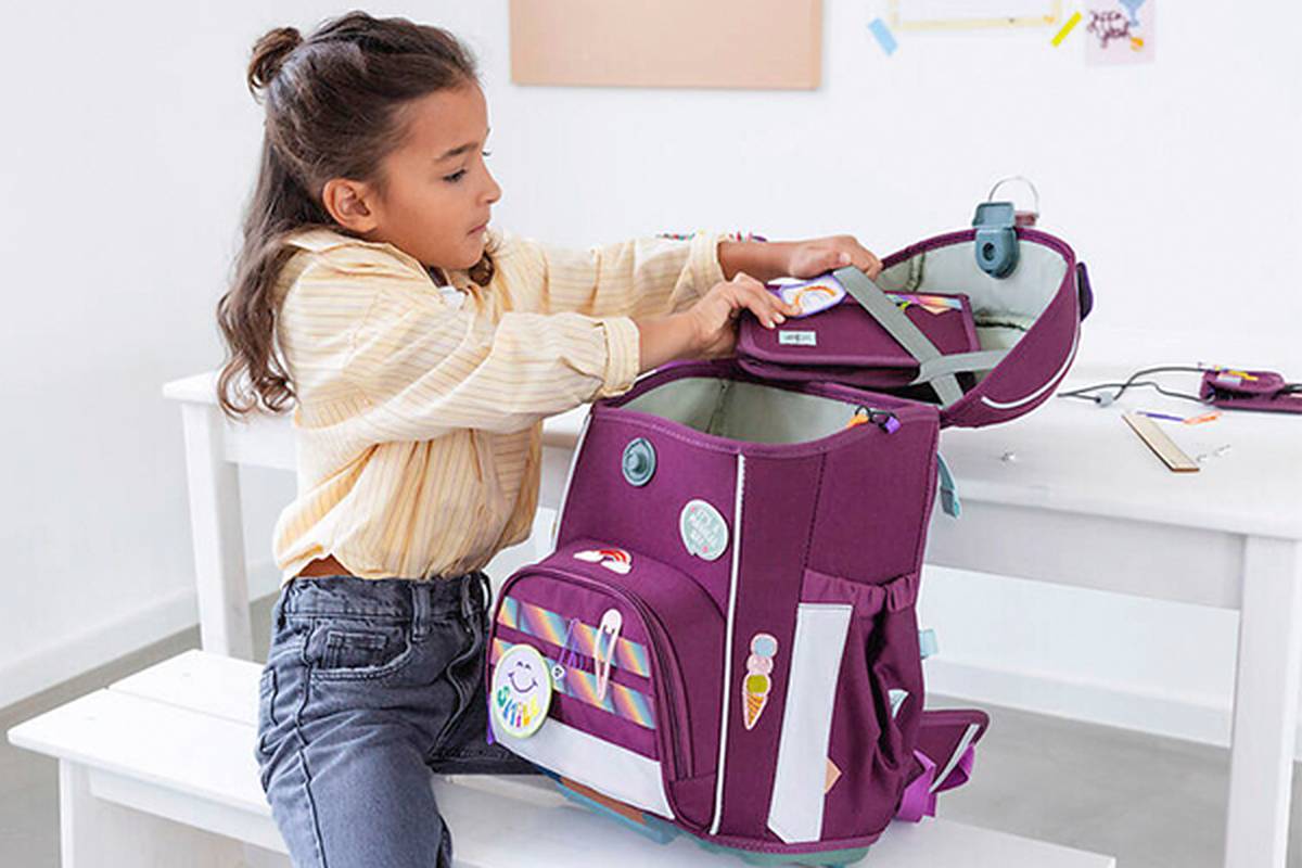 Ein junges Mädchen packt auf einer weißen Bank in einem hellen Raum einen bunten Rucksack mit Schulsachen.