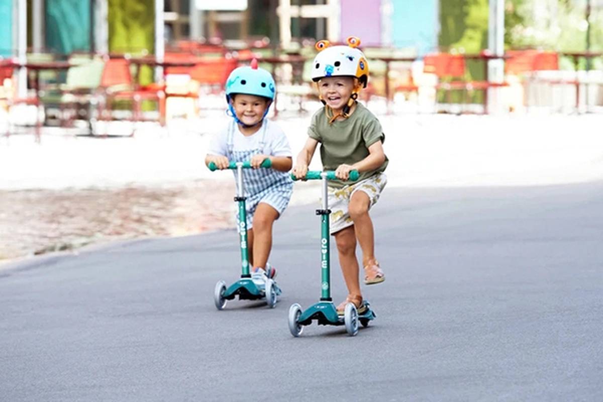 Zwei Kinder mit Helmen fahren fröhlich Scooter auf einer gepflasterten Fläche, mit unscharfen, farbenfrohen Hintergründen aus Stühlen und Gebäuden.