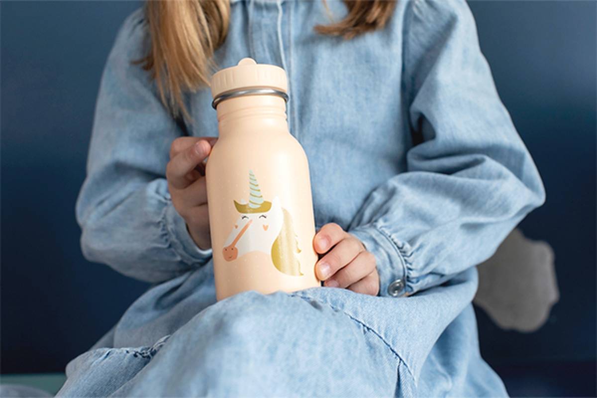 Kind in einem Jeanskleid, das eine pfirsichfarbene Wasserflasche mit einer Einhornabbildung hält.