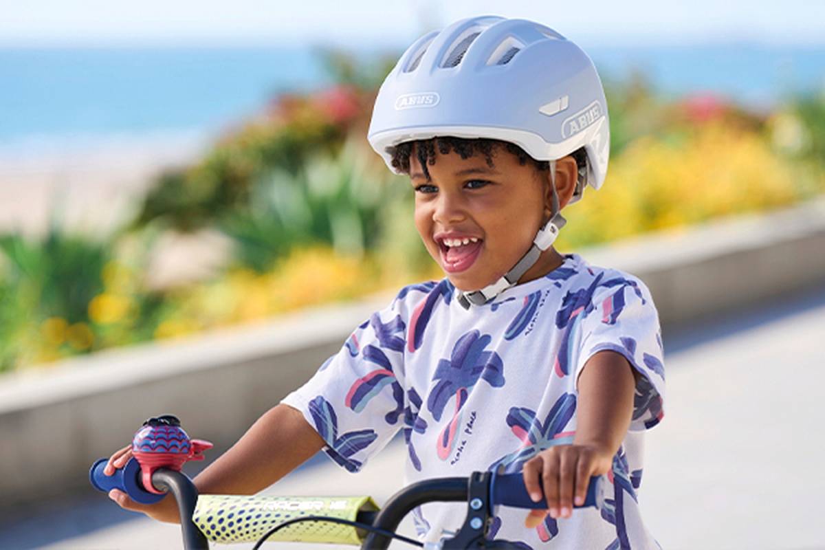Ein Kind mit Helm lächelt, während es an einem sonnigen Tag am Meer Fahrrad fährt.