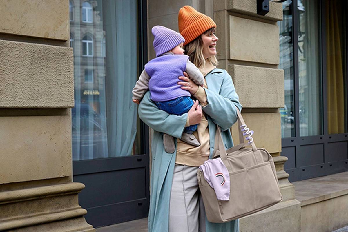 Eine Frau in einem blauen Mantel und einer orangefarbenen Mütze hält lächelnd ein Baby in Lila und Rosa vor einem Gebäude; sie hat eine Wickeltasche über der Schulter.